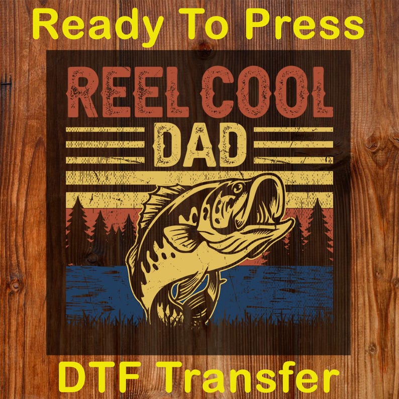 May include: A DTF transfer with a fishing theme. The design features the text "REEL COOL DAD" in red and yellow, with a fish illustration. The background includes a sunset and water scene. The words "Ready To Press" and "DTF Transfer" are also present.