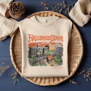 May include: Cream-colored sweatshirt featuring a Halloween-themed graphic. The graphic includes the text "Halloweentown" above a vintage illustration of a town scene with a pumpkin, buildings, and the text "EST. 1998".
