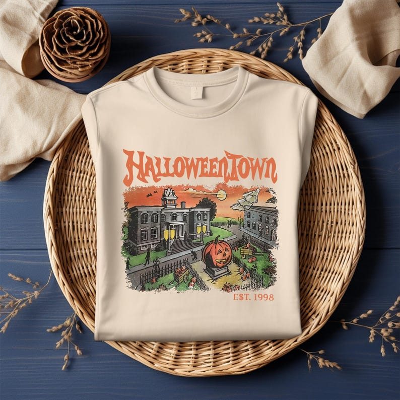 May include: Cream-colored sweatshirt featuring a Halloween-themed graphic. The graphic includes the text "Halloweentown" above a vintage illustration of a town scene with a pumpkin, buildings, and the text "EST. 1998".