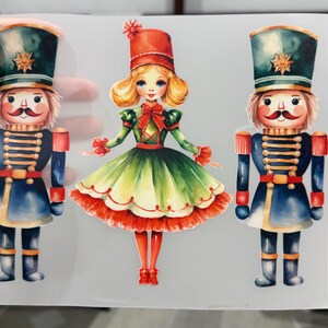 Op de afbeelding: Waterverfillustratie van drie figuren met kerstthema. Twee notenkrakersoldaten in blauwe en rode uniformen flankeren een meisje in een groene en rode jurk. De notenkrakers dragen hoge hoeden met gouden sterren.