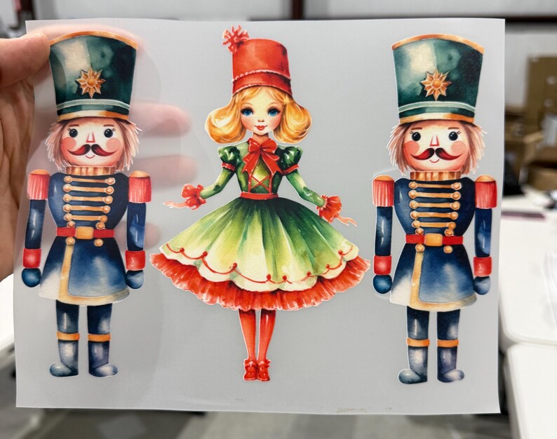Op de afbeelding: Waterverfillustratie van drie figuren met kerstthema. Twee notenkrakersoldaten in blauwe en rode uniformen flankeren een meisje in een groene en rode jurk. De notenkrakers dragen hoge hoeden met gouden sterren.
