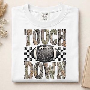 Pode incluir: Uma t-shirt branca de manga curta com uma impress&atilde;o gr&aacute;fica com o texto TOUCH DOWN num padr&atilde;o de folhas de camuflagem. Uma bola de futebol americano preta com atacadores brancos e textura rachada est&aacute; centrada entre as palavras, sobre um fundo axadrezado preto e branco.
