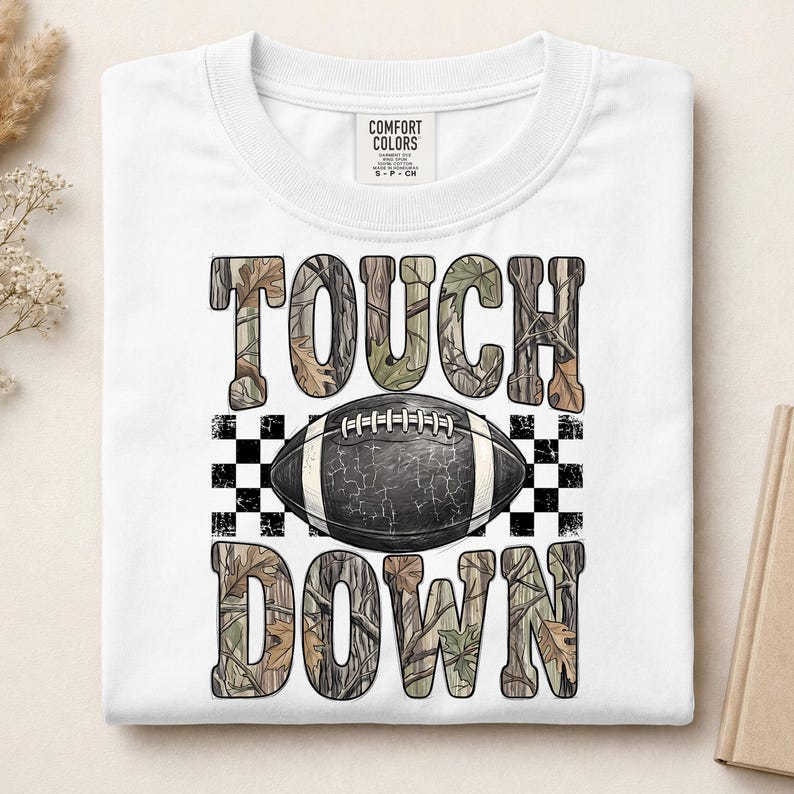 Pode incluir: Uma t-shirt branca de manga curta com uma impress&atilde;o gr&aacute;fica com o texto TOUCH DOWN num padr&atilde;o de folhas de camuflagem. Uma bola de futebol americano preta com atacadores brancos e textura rachada est&aacute; centrada entre as palavras, sobre um fundo axadrezado preto e branco.