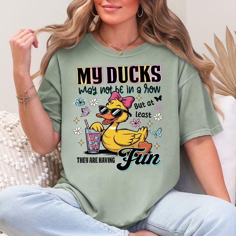 May include: Sage green t-shirt featuring a cartoon duck wearing sunglasses and a pink bow, holding a drink with a straw. The shirt has the text "MY DUCKS may not be in a row But at least THEY ARE HAVING Fun" with floral and butterfly accents.
