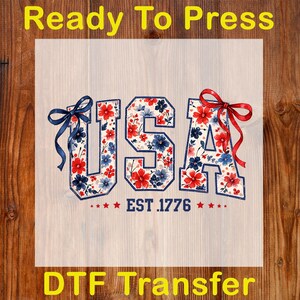 Może przedstawiać: Transfer DTF z napisem "USA" w kwiatowym wzorze z czerwonych, białych i niebieskich kwiat&oacute;w. Litery są obrysowane granatowym kolorem i ozdobione kokardkami. Tekst "EST. 1776" znajduje się pod literami, z gwiazdkami po obu stronach. Obecne są r&oacute;wnież słowa "Ready To Press" i "DTF Transfer".