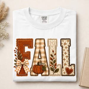 May include: A white short-sleeved t-shirt featuring the word FALL in large block letters. Each letter has a unique texture: the F is solid orange with a plaid bow, the A is beige plaid with a pumpkin, the L is cream cable knit with leaves, and the L is floral with a heart.