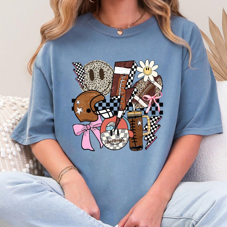 Peut inclure: T-shirt bleu clair avec un motif graphique sur le th&egrave;me du football. Le motif comprend un ballon de football, un casque, une marguerite, une tasse &agrave; caf&eacute; et des &eacute;clairs, tous avec un design r&eacute;tro de visage souriant. Le t-shirt a une coupe d&eacute;contract&eacute;e.