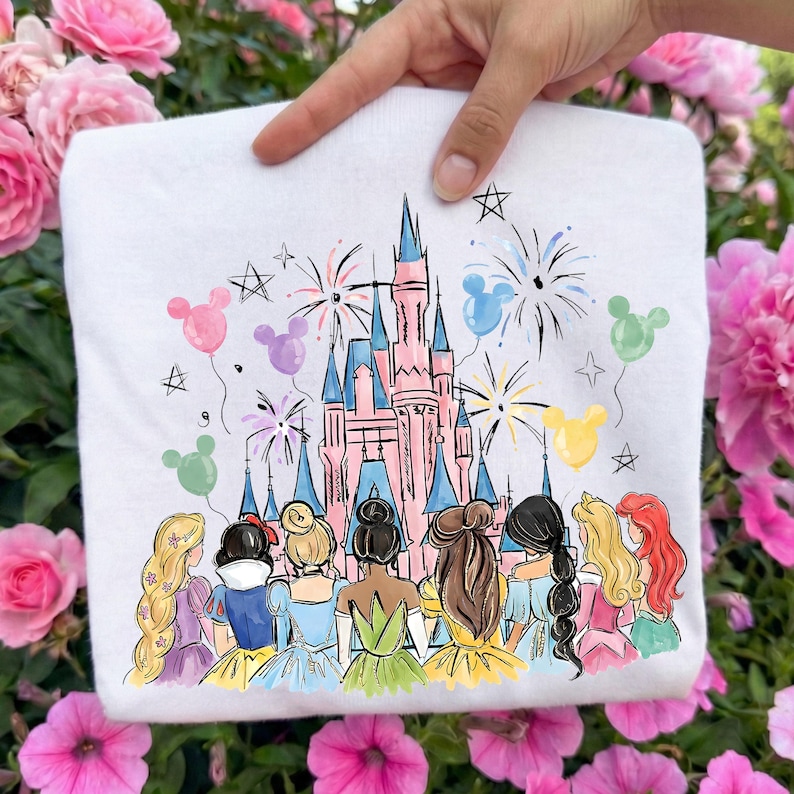 以下が含まれることがあります： ピンクの城、花火、ディズニーのプリンセスキャラクターのカラフルなイラストが描かれた白いスウェットシャツ。ミッキーマウスの風船と星のアクセントがデザインに含まれており、気まぐれで祝祭的なテーマを作り出しています。