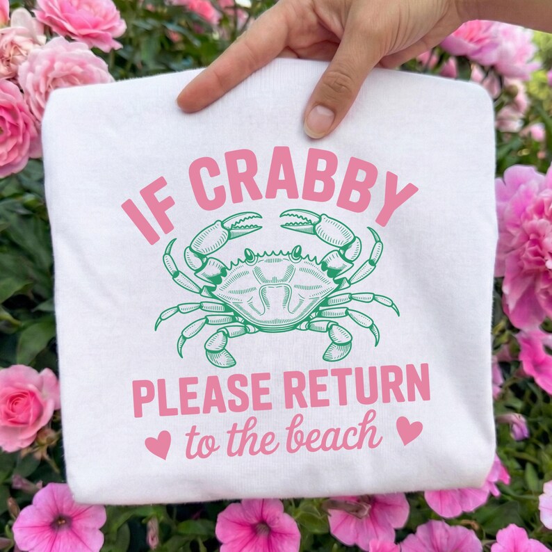 Puede incluir: Sudadera blanca con el texto "IF CRABBY PLEASE RETURN to the beach" en rosa, con una ilustraci&oacute;n de un cangrejo verde. La sudadera es sostenida por una mano, rodeada de flores rosas.