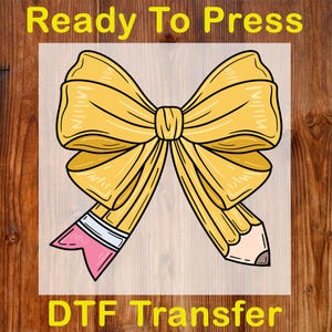 May include: A yellow bow design with a pencil on each side, with a pink eraser and a pencil tip. The text "Ready To Press" is at the top, and "DTF Transfer" is at the bottom.