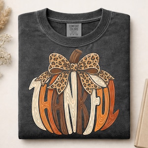 May include: A charcoal gray t-shirt with a graphic of a pumpkin formed by the word THANKFUL in a wood grain texture. The pumpkin is topped with a large leopard print bow. The inner neck label reads COMFORT COLORS, 100% COTTON, S-P-CH.