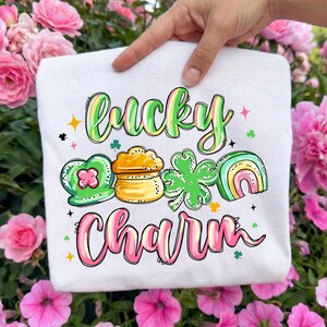 May include: White t-shirt with the words "Lucky Charm" in a colorful, whimsical font. The design includes a green heart-shaped charm, a pot of gold, a four-leaf clover, and a rainbow. The shirt is held up by a hand.