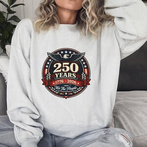 Peut inclure: Un sweat-shirt gris clair avec un motif circulaire comm&eacute;morant 250 ans. Le design comprend un aigle, des &eacute;toiles et le texte "250 YEARS 1776-2026 We The People" en rouge, blanc et bleu.