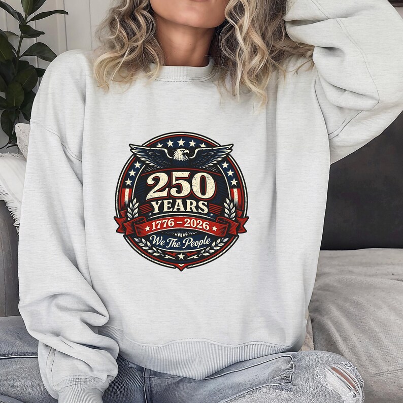 Peut inclure: Un sweat-shirt gris clair avec un motif circulaire comm&eacute;morant 250 ans. Le design comprend un aigle, des &eacute;toiles et le texte "250 YEARS 1776-2026 We The People" en rouge, blanc et bleu.