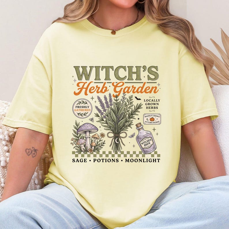 May include: A pale yellow t-shirt featuring a graphic print with the text "WITCH'S Herb Garden". The design includes a central bundle of herbs, mushrooms, a bottle labeled "WITCHES BREW", and text reading "FRESHLY GATHERED", "LOCALLY GROWN HERBS", and "SAGE &bull; POTIONS &bull; MOONLIGHT".