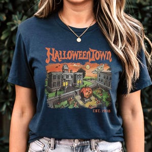 May include: Navy blue t-shirt with a Halloween-themed graphic. The design features the text "Halloweentown" above a spooky town scene with a jack-o'-lantern, buildings, and the text "EST. 1998".