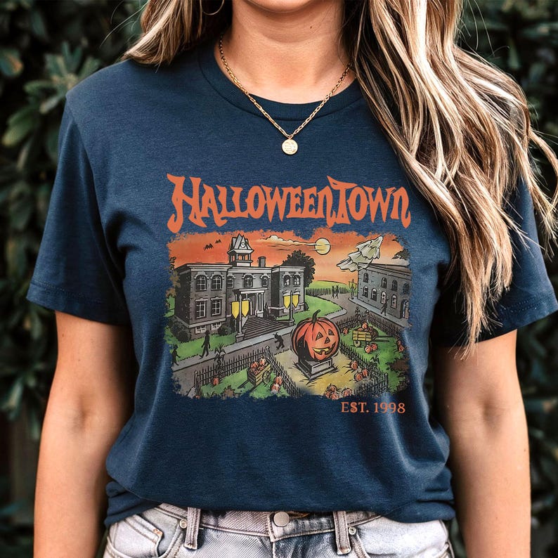 May include: Navy blue t-shirt with a Halloween-themed graphic. The design features the text "Halloweentown" above a spooky town scene with a jack-o'-lantern, buildings, and the text "EST. 1998".