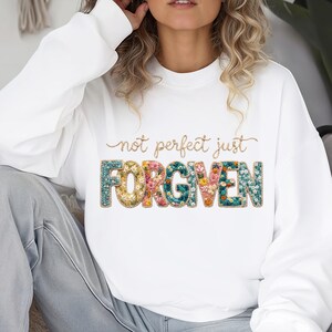 Op de afbeelding: Witte sweatshirt met de tekst "not perfect just FORGIVEN" in gouden letters en bloemrijke letters. Het woord "FORGIVEN" heeft een teal, goud en roze bloemenontwerp. Een casual, comfortabele stijl.