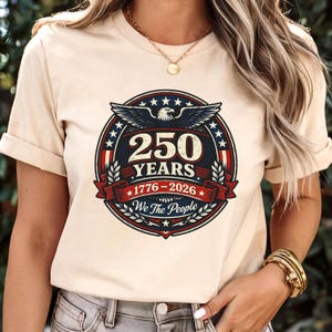 Peut inclure: T-shirt beige avec un motif circulaire comm&eacute;morant 250 ans, de 1776 &agrave; 2026. Le design comprend un aigle, des &eacute;toiles et le texte "We The People" dans un sch&eacute;ma de couleurs patriotiques rouge, blanc et bleu.
