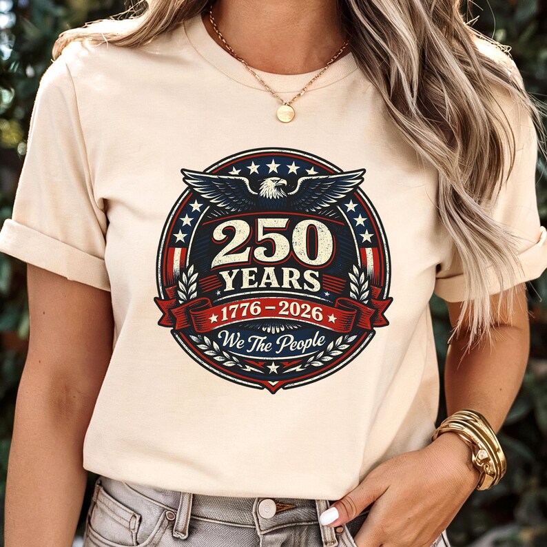 Peut inclure: T-shirt beige avec un motif circulaire comm&eacute;morant 250 ans, de 1776 &agrave; 2026. Le design comprend un aigle, des &eacute;toiles et le texte "We The People" dans un sch&eacute;ma de couleurs patriotiques rouge, blanc et bleu.