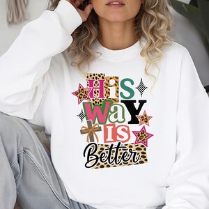 Op de afbeelding: Witte sweatshirt met de tekst "His Way Is Better" in een kleurrijk, gelaagd ontwerp. De tekst bevat luipaardprint, sterren en een kruis. De sweatshirt is een comfortabele, casual stijl.