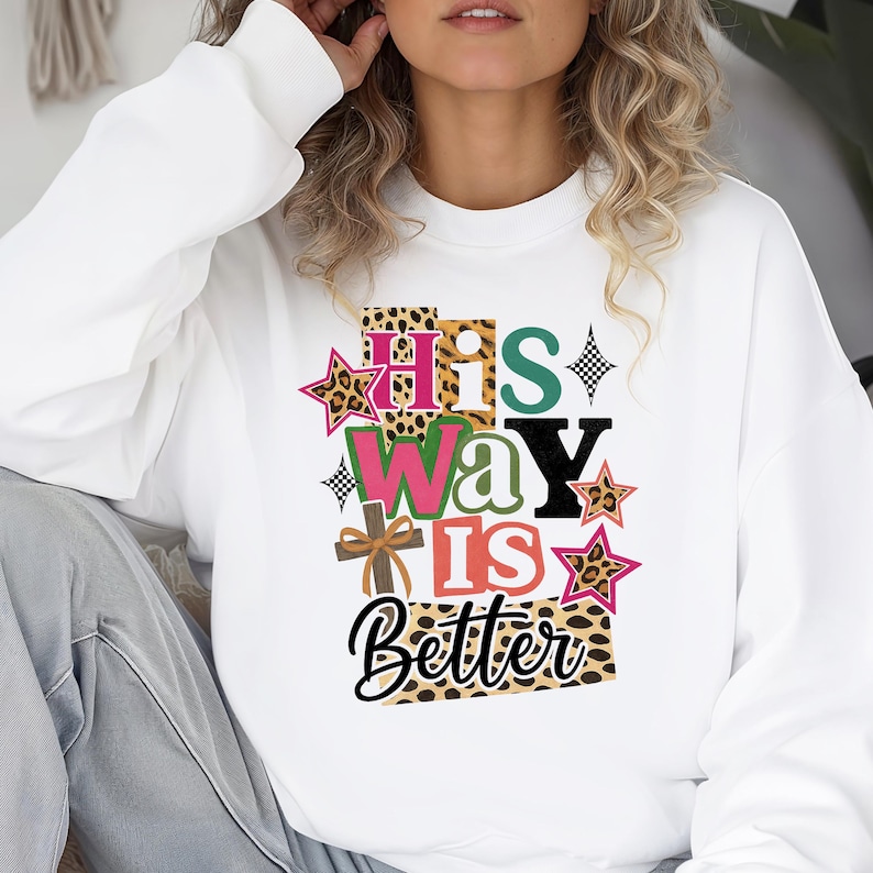 Op de afbeelding: Witte sweatshirt met de tekst "His Way Is Better" in een kleurrijk, gelaagd ontwerp. De tekst bevat luipaardprint, sterren en een kruis. De sweatshirt is een comfortabele, casual stijl.