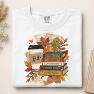 May include: A white t-shirt featuring a graphic print of a coffee cup with a sleeve labeled fall, stacked books titled Crisp Air, Cozy Days, Autumn Stories, Pumpkin Spice, and Sweater Weather, surrounded by autumn leaves and vintage paper scraps.
