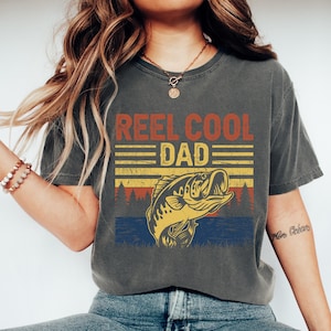 May include: Dark gray t-shirt with the text "REEL COOL DAD" in red and yellow. Below the text is a graphic of a fish jumping out of the water, with a forest silhouette in the background. The shirt is a casual style.