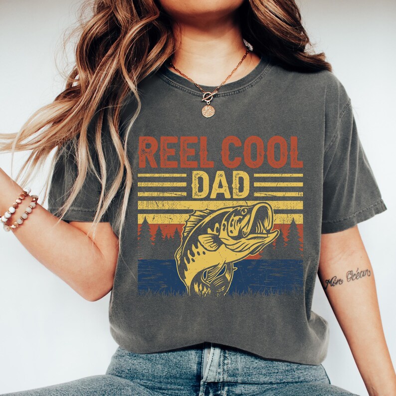May include: Dark gray t-shirt with the text "REEL COOL DAD" in red and yellow. Below the text is a graphic of a fish jumping out of the water, with a forest silhouette in the background. The shirt is a casual style.