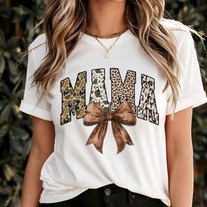 May include: White t-shirt with the word "MAMA" in a bold, patterned design. The letters feature camouflage, animal print, and a cowhide pattern. A large brown bow is centered below the word, adding a decorative element to the shirt.
