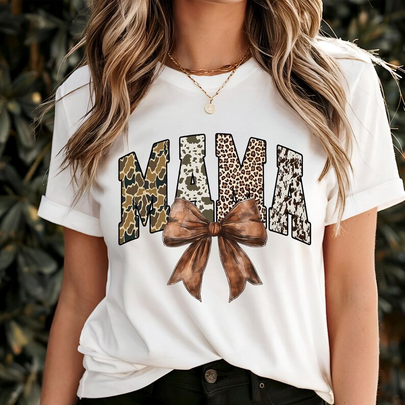 May include: White t-shirt with the word "MAMA" in a bold, patterned design. The letters feature camouflage, animal print, and a cowhide pattern. A large brown bow is centered below the word, adding a decorative element to the shirt.