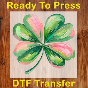 May include: A four-leaf clover design with green and pink hues, ready to press. The image features the text "Ready To Press" at the top and "DTF Transfer" at the bottom, set against a wood grain background.