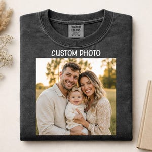 May include: A charcoal gray crew neck t-shirt featuring a rectangular custom photo print of a family consisting of an adult, a baby, and another adult. The shirt has a Comfort Colors brand label at the neck with size S-P-CH and text CUSTOM PHOTO.