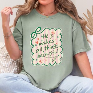 May include: Sage green t-shirt with the text "He makes all things beautiful" in a decorative frame. The design includes hearts, stars, and floral elements. The text "Ecclesiastes 3:16" is also included.