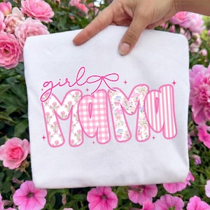 May include: A white sweatshirt with the words "girl mama" in pink. The word "girl" is in cursive, and "mama" is in a floral, gingham, and striped pattern. The sweatshirt is held up by a hand, surrounded by pink flowers.