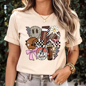 Peut inclure: T-shirt beige avec un collage graphique sur le th&egrave;me du football. Le motif comprend un ballon de football, un casque, un smiley, des &eacute;clairs et une marguerite, le tout sur un fond &agrave; carreaux. Un n&oelig;ud rose ajoute une touche de couleur.
