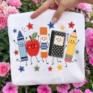 May include: A white t-shirt featuring an embroidered design of five anthropomorphic school supplies: a blue gingham crayon, a red plaid apple, an orange glue stick, a black polka-dot notebook, and a yellow plaid pencil, surrounded by colorful stars.