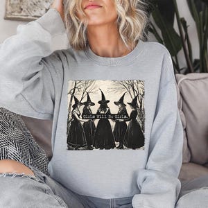 Puede incluir: Sudadera gris claro con un estampado en blanco y negro de cinco brujas con vestidos y sombreros negros, con el texto "Girls Will Be Girls". La ilustraci&oacute;n est&aacute; sobre un fondo de &aacute;rboles desnudos.