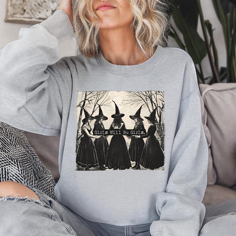 Puede incluir: Sudadera gris claro con un estampado en blanco y negro de cinco brujas con vestidos y sombreros negros, con el texto "Girls Will Be Girls". La ilustraci&oacute;n est&aacute; sobre un fondo de &aacute;rboles desnudos.