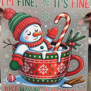 Peut inclure: Illustration d'un bonhomme de neige dans un bonnet, une &eacute;charpe et des moufles rouges et vertes, assis dans une tasse rouge. La tasse pr&eacute;sente des motifs de flocons de neige et le texte "I'M FINE, IT'S FINE, JUST HAVING A MELTDOWN". Une canne en bonbon et des b&acirc;tons de cannelle sont &eacute;galement pr&eacute;sents.