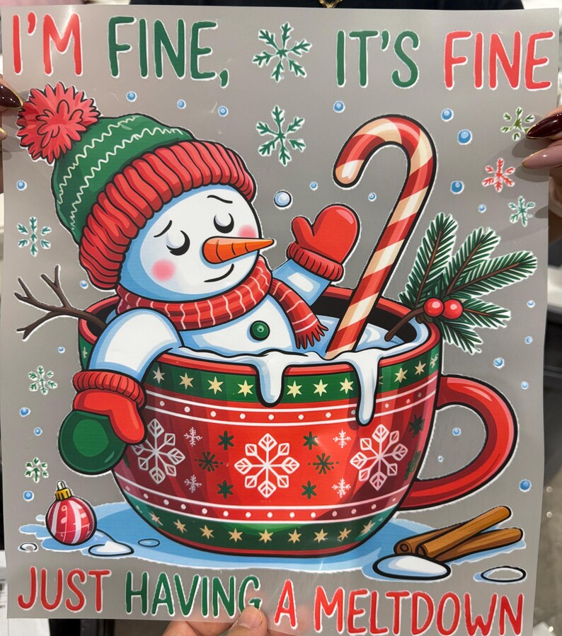 Peut inclure: Illustration d'un bonhomme de neige dans un bonnet, une &eacute;charpe et des moufles rouges et vertes, assis dans une tasse rouge. La tasse pr&eacute;sente des motifs de flocons de neige et le texte "I'M FINE, IT'S FINE, JUST HAVING A MELTDOWN". Une canne en bonbon et des b&acirc;tons de cannelle sont &eacute;galement pr&eacute;sents.