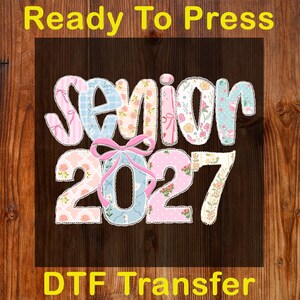 Peut inclure: Un motif de transfert DTF avec les mots "Ready To Press" et "DTF Transfer" en jaune. Le motif pr&eacute;sente le mot "Senior" et le chiffre "2027" dans un motif floral et pastel, avec un n&oelig;ud rose.