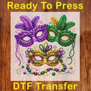 May include: A DTF transfer featuring three ornate Mardi Gras masks in purple, green, and gold. The masks are decorated with feathers, beads, and intricate designs. The text "Ready To Press" and "DTF Transfer" are displayed in yellow.