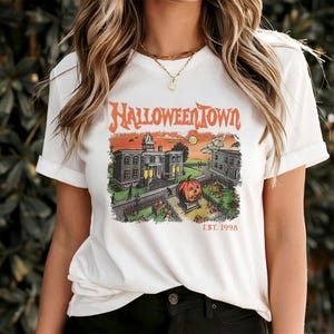 May include: White t-shirt with a HalloweenTown graphic. The graphic features a vintage-style illustration of a town scene with a jack-o'-lantern, buildings, and the text "HalloweenTown EST. 1998" in orange.