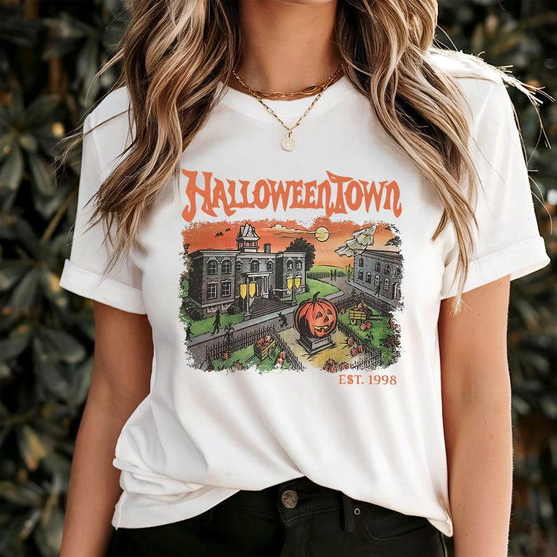 May include: White t-shirt with a HalloweenTown graphic. The graphic features a vintage-style illustration of a town scene with a jack-o'-lantern, buildings, and the text "HalloweenTown EST. 1998" in orange.