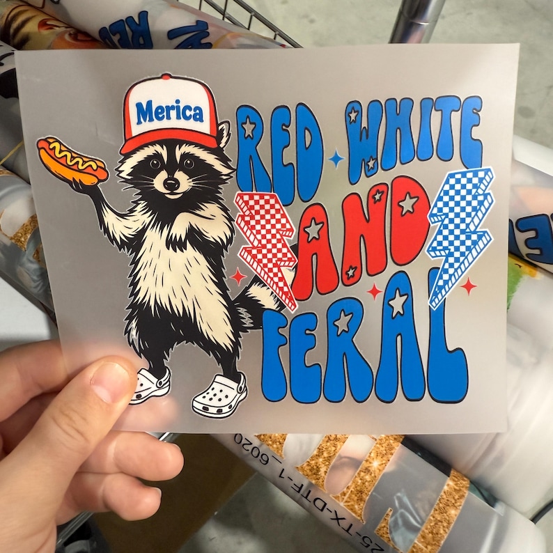 May include: A transparent graphic featuring a raccoon wearing a hat that says "Merica" and holding a hot dog. The text "RED WHITE AND FERAL" is in blue and red, with lightning bolt graphics.