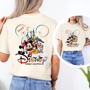 May include: Cream-colored t-shirt featuring a colorful Disney family vacation design. The graphic includes Mickey and Minnie Mouse, Donald Duck, Goofy, and other characters, with the text "2026" and "Disney Family Vacation".