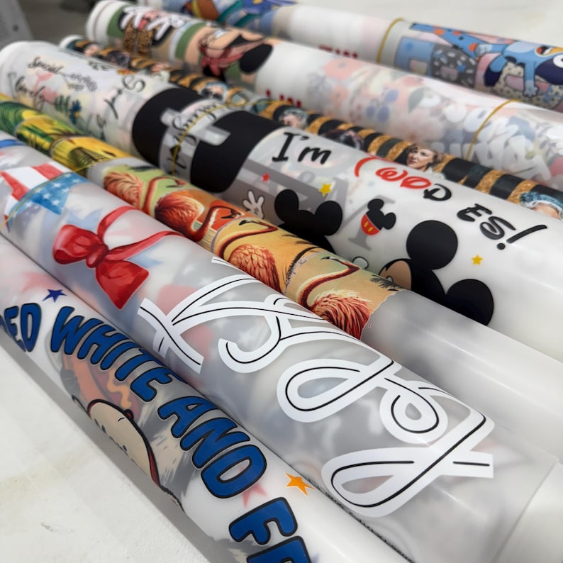 May include: Several rolls of printed fabric are displayed, showcasing various designs. One roll features a red bow and American flag imagery, while others display cartoon characters and text like "I'm" and "Red, White, and Free". The rolls are tightly wound.