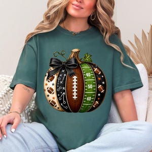 May include: A dark green t-shirt featuring a graphic of a pumpkin designed with American football elements. The pumpkin has segments showing a football field, a brown football texture, and black sections with white play diagrams and stars.