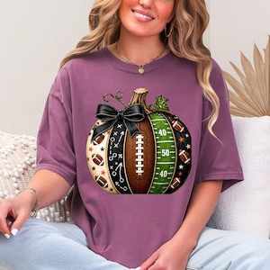 May include: A mauve short-sleeve t-shirt featuring a graphic of a pumpkin designed with football elements. The pumpkin includes a black bow, a brown football texture, a green field pattern with yard lines, and small football and star icons.